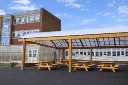 Timber-framed barrel-vaulted dining canopy with picnic benches at Blatchington Mill High School, providing a covered outdoor seating area beside a brick school building.