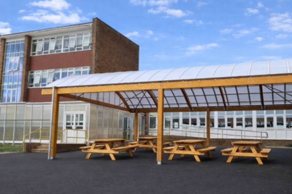 Timber-framed barrel-vaulted dining canopy with picnic benches at Blatchington Mill High School, providing a covered outdoor seating area beside a brick school building.