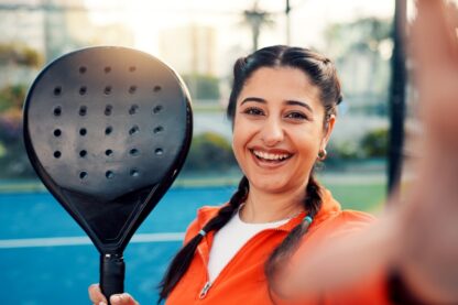 Smiling woman holding a padel racket on an outdoor court, wearing an orange jacket and looking at the camera.