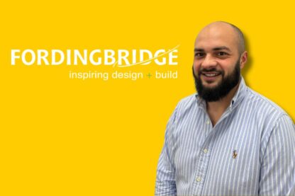 Man smiling in front of a yellow Fordingbridge background with “inspiring design + build” text, wearing a striped shirt.