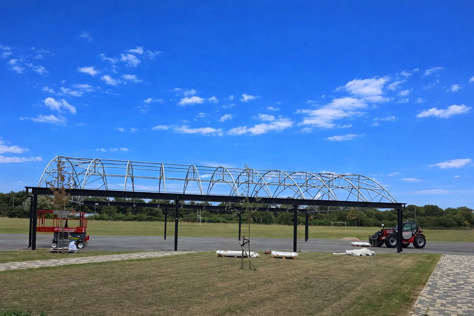 Invite team members Uploaded image can you please make the sky nice and blue? Generated image: Construction site under bright blue sky Edit can I please have this as a jpg Here you go: [Download the JPG](sandbox:/mnt/data/blue can you please provide the ALT text for this image A long canopy structure under construction on a school field, with metal framework supported by black columns, construction equipment on either side, and a bright blue sky with scattered clouds overhead.