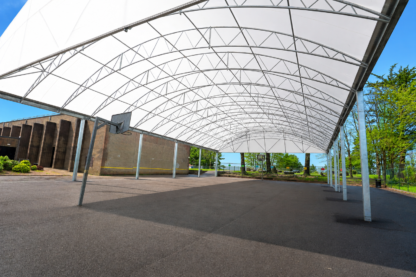 Large white tensile canopy at Saltash Community School creating a covered outdoor sports and social space, with steel arch framework, basketball hoop and open hard-surfaced area beneath on a bright sunny day.