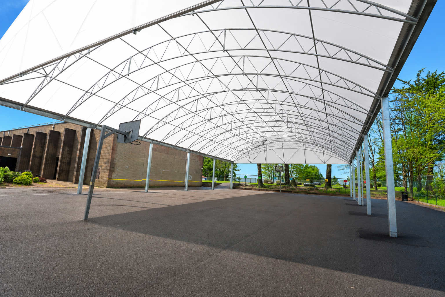 Large white tensile canopy at Saltash Community School creating a covered outdoor sports and social space, with steel arch framework, basketball hoop and open hard-surfaced area beneath on a bright sunny day.