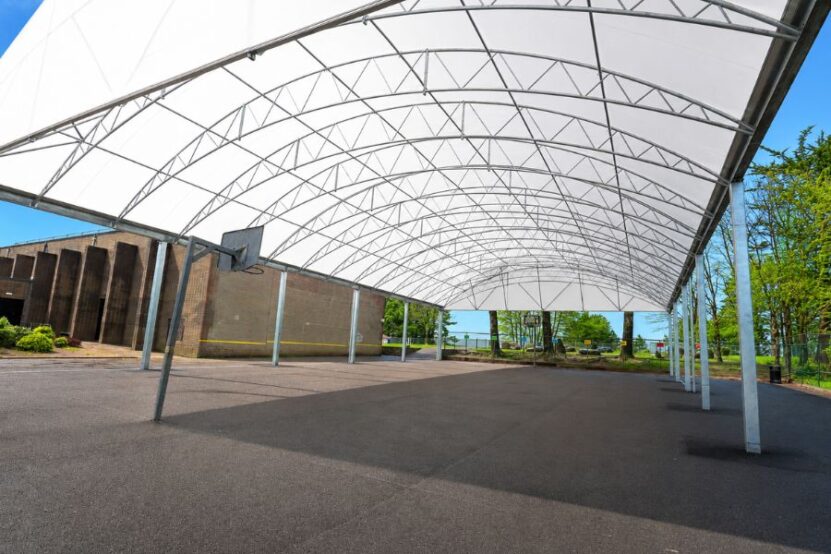 Large all-weather school canopy providing shade and weather protection over an outdoor games and learning area on a bright summer day.